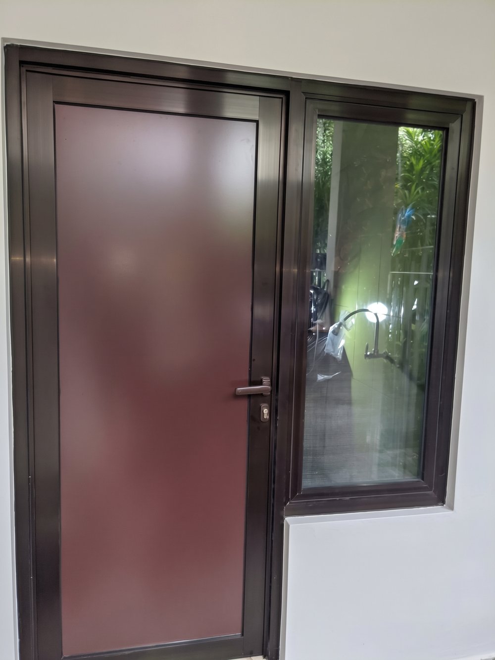 Pintu ACP swing warna marun dengan rangka aluminium coklat dan jendela kaca mati di sampingnya